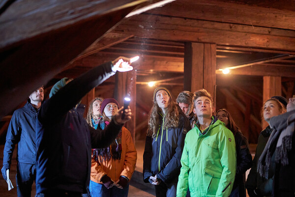 Freiwillige stehen in einem Holzbau und schauen an die Decke, die mit einer Taschenlampe angeleuchtet wird.